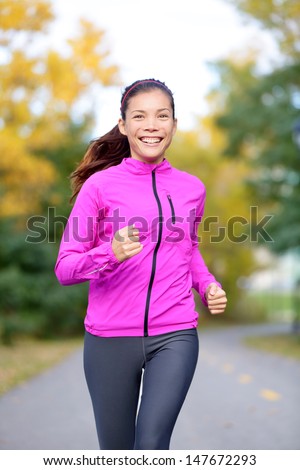 Running sport woman training in fall autumn forest or city park smiling happy. Asian female jogger working out outdoor energetic and fresh. Mixed race Asian Caucasian female fitness model.