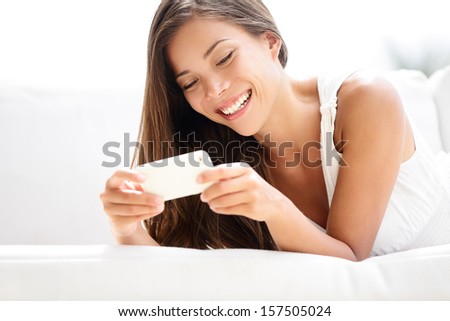 Smartphone Woman Using App On Mobile Smart Phone Smiling Happy. Beautiful Multiracial Girl Sms Text Messaging Or Using Application While Lying On Sofa. Asian Caucasian Model In Her 20s.