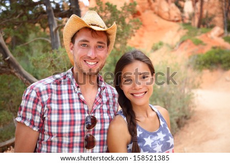 Happy outdoors couple portrait in american countryside. Smiling multiracial young couple in western USA nature. Man wearing cowboy hat and woman wearing USA flag shirt.