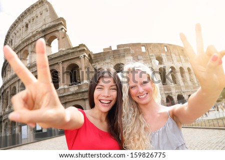 Travel tourist girl friends by Colosseum, Rome. Happy girlfriends tourists showing victory hand sign gesture in front of Coliseum. Beautiful young happy blonde girl and multiracial Asian woman, Italy.