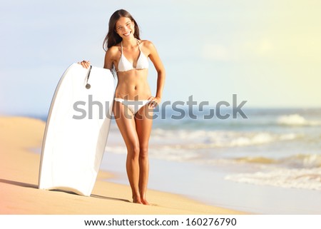 Surfer bikini woman on beach smiling with surfboard smiling holding body board. Pretty smiling girl in white bikini standing on beach enjoying summer sun. Mixed race Asian Caucasian, Maui, Hawaii, USA