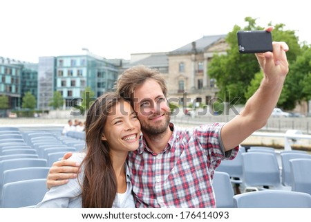 Tourist Couple On Travel In Berlin, Germany On Boat Tour Cruise Smiling Happy Taking Selfie Self-Portrait Photo Picture While Enjoying Their Romantic Europe Travel Vacation. Asian Woman, Caucasian Man