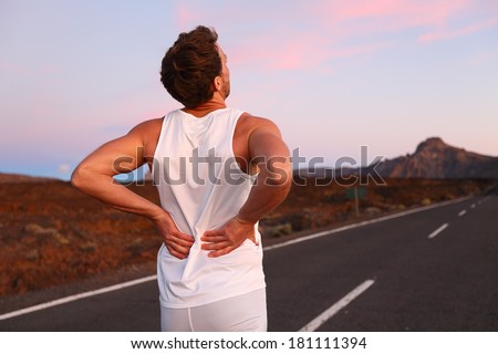 Back pain. Athletic running man with injury in sportswear rubbing touching lower back muscles standing on road outside at night.