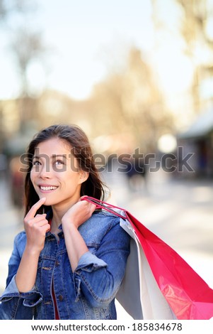 Shopping woman thinking looking up outdoors in fashion jeans jacket. Shopper girl holding shopping bags up on walking street. Mixed race Asian Caucasian female model La Rambla street Barcelona.