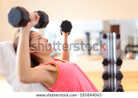 Gym woman strength training lifting dumbbell weights in shoulder press exercise. Female fitness girl exercising indoor in fitness center. Beautiful fit mixed race Asian Caucasian model training.