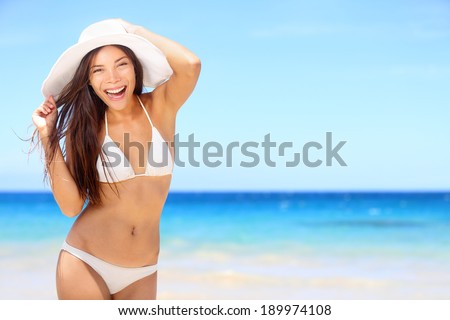 Beach woman happy on travel vacation holidays in bikini on by blue ocean sea at tropical resort. Cheerful smiling excited mixed race girl wearing sun hat laughing full of joy looking at camera.