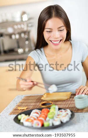Young pretty asian mixed race asian caucasian model taking healthy lifestyle sushi from plate - about to eat happily smiling looking at food. Sitting at table in kitchen