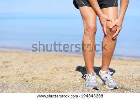 Running injury - Man jogging with knee pain. Close-up view of runner injured jogging on the beach clutching his knee in pain. Male fitness athlete.