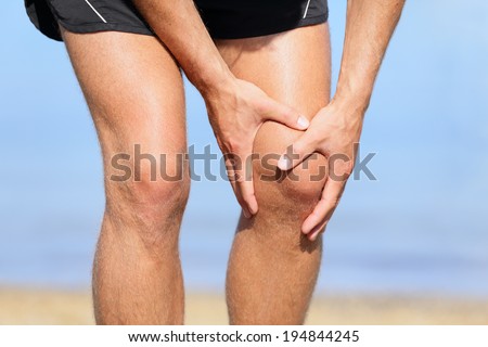 Runner injury - Man running with knee pain. Close-up view of runner injured jogging on the beach clutching his knee in pain. Male fitness athlete.