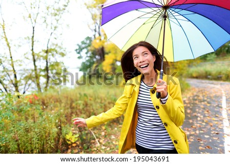 Autumn woman happy in rain running with umbrella. Female model looking up at clearing sky joyful on rainy fall day wearing yellow raincoat outside in nature forest by lake. Multi-ethnic Asian girl.