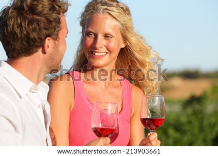 Happy couple drinking wine in sunset. Red or rose glasses at vineyard. Romantic couple enjoying glass of wine smiling happy in love outdoors. Young Caucasian blonde woman in focus.
