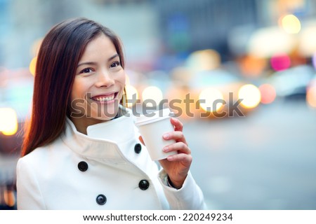 Young casual urban professional woman in New York City Manhattan drinking coffee walking in street wearing coat downtown with yellow taxi cabs in background. Female business woman.