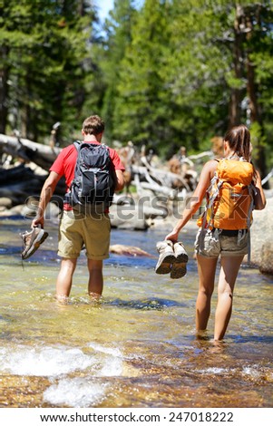 Hikers group walking barefoot crossing river in forest. Adventure people on hike hiking in nature holding shoes and boots to cross with wet feet.