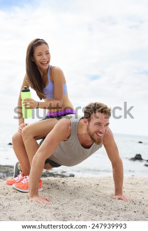 Fitness couple training doing funny push-up on beach during workout. Woman playful having fun sitting on boyfriend to test his strength with heavy weight.