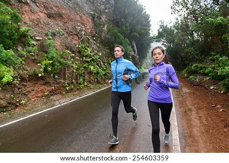 Healthy lifestyle people running on country road exercising. Runners jogging on mountain road training for marathon. Asian woman and Caucasian man wearing waterproof sports clothing for wind and rain.