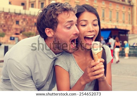 Funny playful young couple eating ice cream. Goofy portrait of boyfriend teasing girlfriend biting off a cold dessert in city summer, Stockholm, Sweden, Europe.