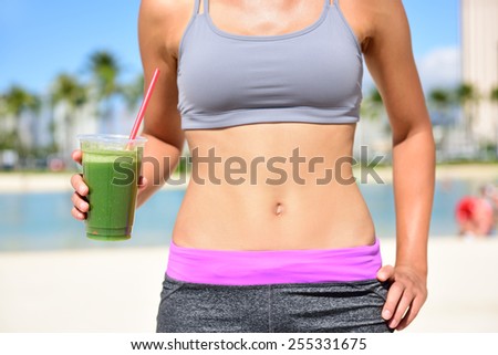 Healthy lifestyle fitness woman drinking green vegetable smoothie juice after running exercise.  Close up of smoothie and stomach. Healthy lifestyle concept with fit female model outside on beach.