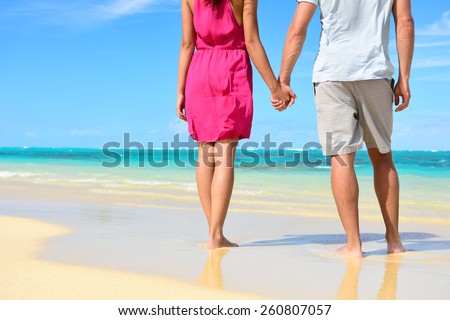 Beach couple in love holding hands on honeymoon. Lower body crop showing pink dress, casual beachwear, legs and feet of romantic newlyweds people standing on white sand on travel summer vacations.