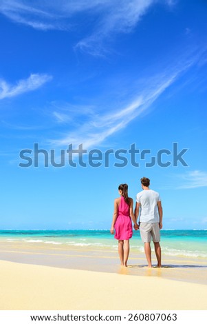 Beach vacation couple relaxing on summer holidays. Young people standing from behind holding hands looking at the ocean, vertical crop with a lot of copy-space in the blue sky. Travel concept.
