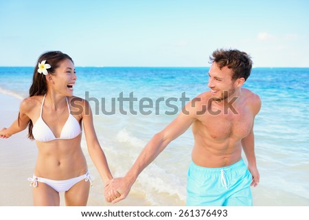 Beach couple having fun happy on beach vacation during summer holiday. Multiracial fit couple running together holding hands laughing in the sun. Young adults in shape carefree happiness.
