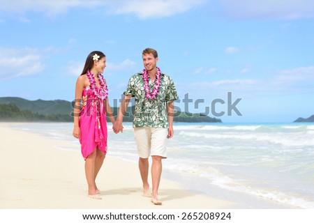 Happy couple on Hawaii vacation walking on beach with Hawaiian leis and Aloha clothing. Caucasian man wearing typical Hawaiian shirt and Asian woman girlfriend in pink sarong fabric sundress.