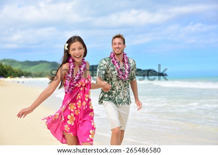Happy couple having fun running on Hawaii beach vacations in Hawaiian clothing wearing Aloha shirt and pink sarong sun dress and flower leis for traditional wedding or honeymoon concept.