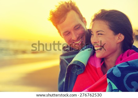 Bathing romantic couple with towel on beach sunset. Portrait of happy young interracial couple embracing each other having fun outdoors during holidays vacation travel. Asian woman, Caucasian man.