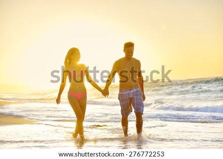 Romantic honeymoon couple in love holding hands walking on beautiful sunset at beach in waterfront. Lovers or newlywed married young couple by the sea enjoying vacation travel holiday. Oahu Hawaii USA