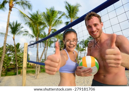 Happy beach volleyball players thumbs up. Excited smiling man and woman with beach volley ball giving thumbs up success hand sign looking at camera. Asian woman, Caucasian man.