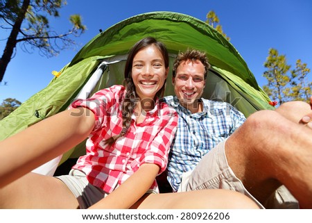 Selfie camping people in tent taking self portrait using camera smartphone. Couple of campers taking picture smiling happy outdoors in forest. Happy people having fun. Asian woman, Caucasian man.