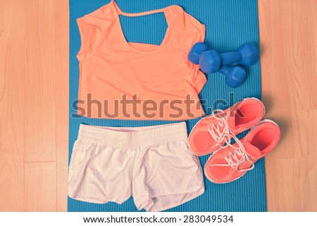 Workout clothes - fitness outfit and running shoes. Overhead of clothing ready for lifting weights at the gym or at home, laying on a yoga mat on the floor. Matching orange t-shirt and sneakers.