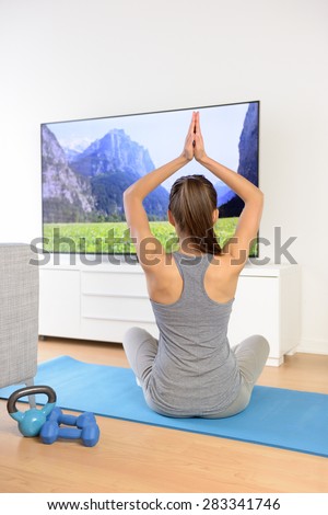 Woman doing home yoga meditation in front of TV. Fit girl doing easy pose relaxation exercises watching a TV show or training video with arms raised sitting on the floor of the living room.