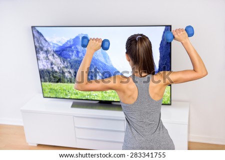 Fitness at home - woman working out in front of tv. Back of a young female adult watching television during her workout, lifting weights to tone arms and shoulders, following an exercise video.
