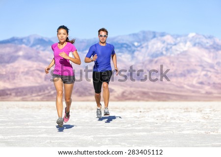 Runners trail running on dry desert landscape. Couple of fit athletes sprinting in compression activewear wearing sports clothing sweating in hot weather. Full length people in dramatic nature.