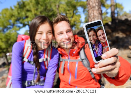 Selfie couple using smart phone camera taking photo hiking in nature with smartphone. Happy couple taking self-portrait picture using app. Man and woman having fun together. Focus on screen.