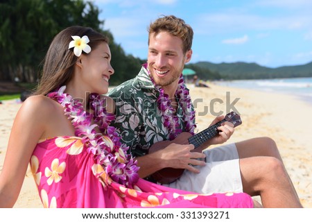 Man on beach playing ukulele instrument on Hawaii with having fun. Young couple, woman and man in love on beach vacations in Hawaiian clothing wearing Aloha shirt dress and flower lei.