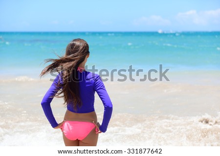 Beach girl going swimming in rashguard swimwear protective clothing. Woman standing in bikini and uv sun protection surf shirt looking at ocean waves. Healthy active lifestyle.