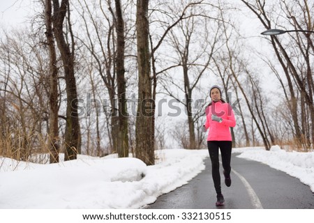 Woman jogging in snowy city park - winter fitness. Female athlete exercising outside in cold weather on forest path wearing activewear. Windbreaker pink jacket, warm tights, running shoes.