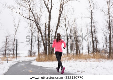 Woman runner running in winter snow - active lifestyle. Female athlete from the back jogging training her cardio on city park path outside in cold weather wearing leggings and coat.
