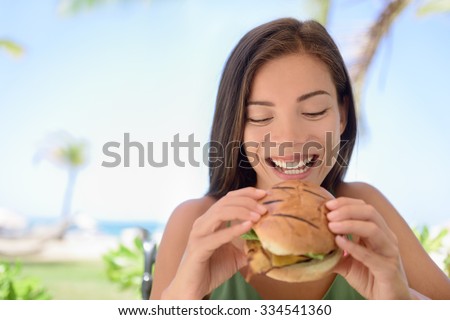 Happy woman holding eating fresh burger sandwich at beach. Young female is sitting at outdoor restaurant. Beautiful tourist is having fresh burger during summer vacation holidays.