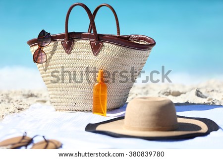 Women's beach accessories on sand for summer vacation concept. Straw tote bag, sun hat and sunscreen lotion or suntan tanning oil spray bottle with blue ocean background for travel holidays.