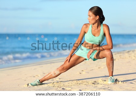 Runner woman stretching legs with lunge hamstring stretch exercise leg stretches. Fitness female athlete relaxing on beach doing a warm-up before her strength training cardio workout.