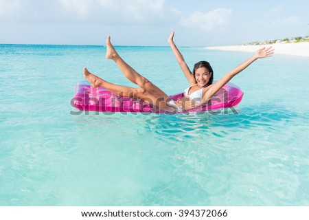 Beach fun girl playful on pink pool float mattress floating on ocean water vacation getaway. Party woman cheering legs and arms up on inflatable toy bed swimming in turquoise sea.