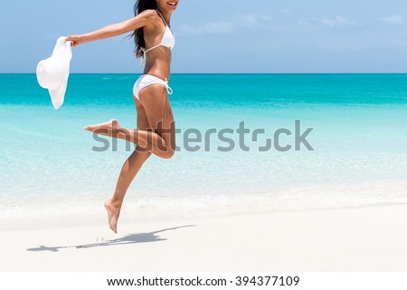 Beach ready bikini body - sexy slim legs and toned thighs and butt. Suntan happy woman jumping in freedom on white sand with sun hat. Weight loss success or epilation concept.