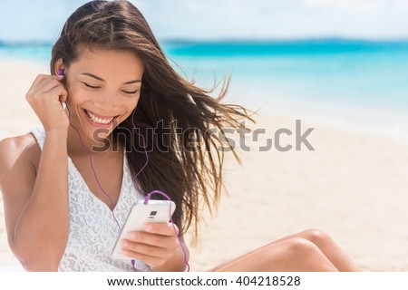 Happy smartphone woman listening with earbuds to streaming music. Young Asian casual girl using her mobile phone app 4g data to play songs while relaxing on beach summer vacations.