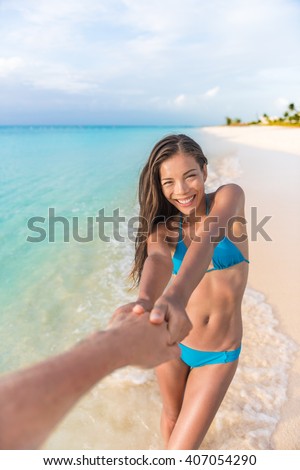 Follow me concept. Asian girlfriend leading man holding hand. Happy multiracial woman in blue bikini with sexy and slim suntan body. Couple in love walking on tropical beach vacations summer travel.