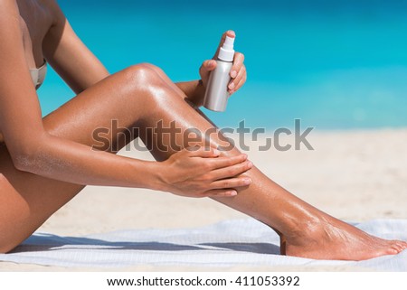 Sunscreen suntan lotion in spray bottle. Young woman in spraying tanning oil on her leg from bottle. Lady is massaging sunscreen lotion while sunbathing at beach. Female model during summer vacation.