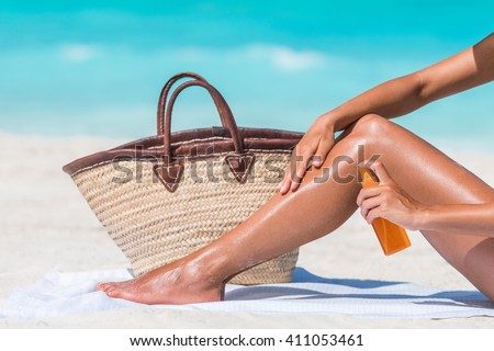 Sunscreen suntan lotion spray bottle woman spraying tanning oil on her leg from bottle. Lady is massaging sunscreen lotion while sunbathing at beach. Unrecognizable model closeup on summer vacation.