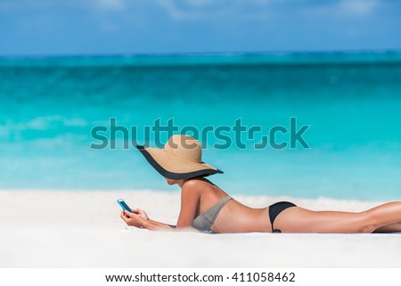Beach vacation girl using mobile phone app texting sms or browsing on online social media during summer travel holiday. Woman relaxing sunbathing on sand lying on towel wearing sun hat.