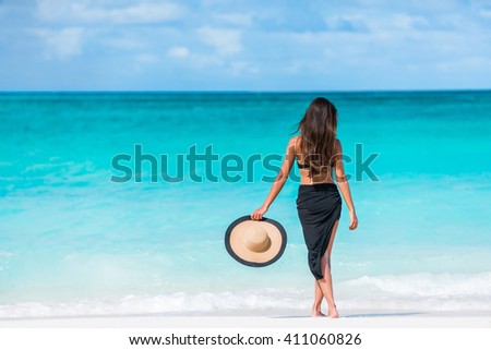Woman in black bikini and sarong standing on beach. Elegant sexy female is wearing black bikini and sarong on beach. Woman is holding sunhat enjoying her summer vacation at resort in the Caribbean.